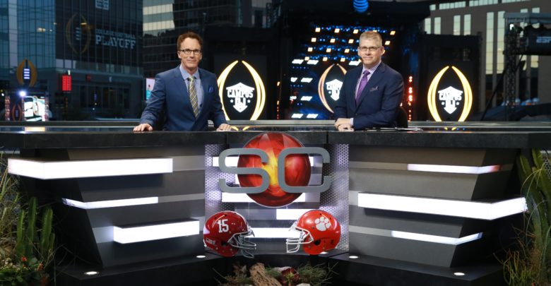 2 men sitting at a table talking about sports with 2 red football helmets in front of the table